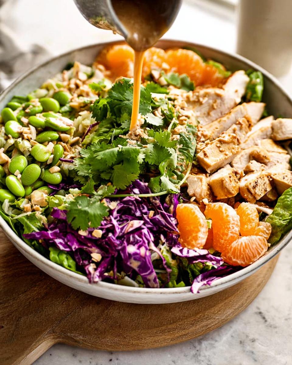 Asian Sesame Chicken Salad with 33g Protein 4 Pouring almond dressing over a vibrant Asian Sesame Chicken Salad with edamame, mandarin oranges, and red cabbage.