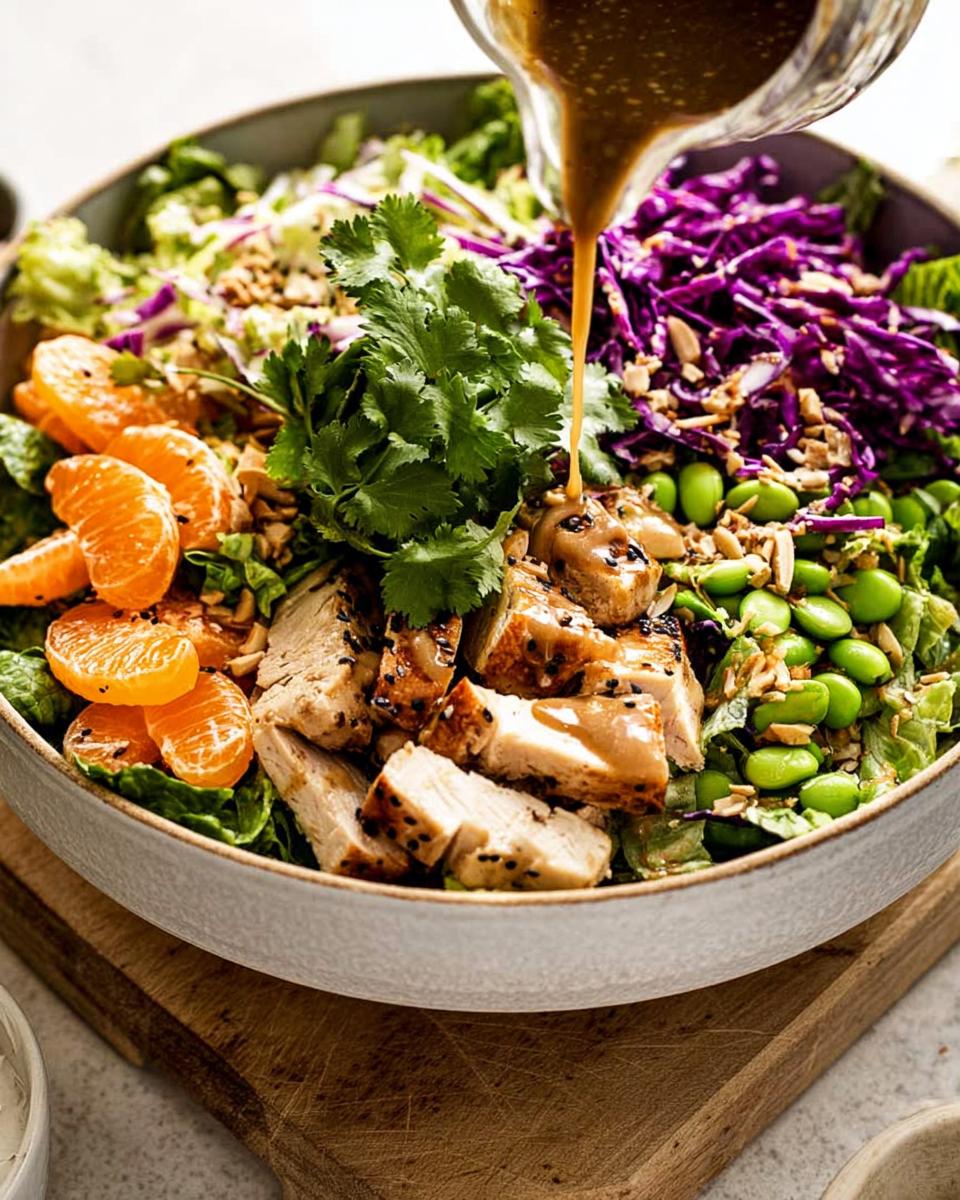 Asian Sesame Chicken Salad with 33g Protein 5 Pouring almond dressing over a vibrant Asian Sesame Chicken Salad with grilled chicken, edamame, mandarin oranges, and red cabbage.
